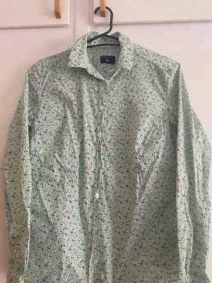 Blommig skjorta från Gant - Snygg blommig skjorta från Gant i vitt med gröna och rosa detaljer. Skjortan har klassisk krage, knappar framtill och lång ärm. Perfekt för dig som vill sticka ut lite extra med ett färgglatt mönster.