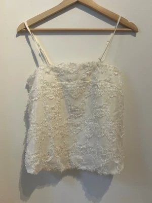 Vit linnetopp med spetsdetaljer - Säljer en ljus, ärmlös linnetopp med tunna axelband och justerbar passform. Toppen har ett unikt spetsmönster med fluffiga detaljer som ger en romantisk och trendig look. Perfekt att styla med jeans eller kjol för en fräsch outfit.