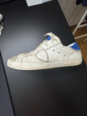 Philippe Model - Säljer ett par vita sneakers från Philippe Model med blå detalj på hälen och blå logga på plösen. Skorna har klassisk låg profil, vita snören och ovandel i läder. Perfekta för dig som gillar stilrena och tidlösa sneakers.