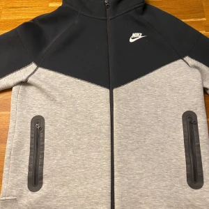 Nike tech - Nike tech i svart och ljusgrått med huva och hel dragkedja. Jackan har två fickor med dragkedjor framtill och en stilren design med Nike-logga på bröstet. Perfekt för dig som gillar sportig och clean stil. Priset kan diskuteras vid snabbaffär.