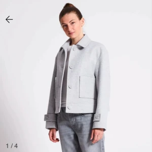 Ljusgrå kappa med knappar - Snygg ljusgrå kappa med knappar framtill och stora fickor. Jackan har en klassisk krage och långa ärmar med dekorativa knappar vid ärmsluten. Perfekt för en stilren look.En knapp på höger arm är sönder. 