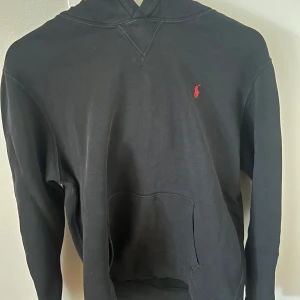 Svart hoodie från Ralph Lauren - Säljer en klassisk svart hoodie från Ralph Lauren med det ikoniska röda logotypbroderiet på bröstet. Tröjan har en känguruficka och en bekväm huva. Perfekt för en avslappnad stil. Priset är inte hugget i sten så hör av er vid minsta fundering!