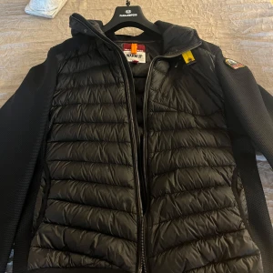 Parajumper cardigan - Säljer en svart jacka från Parajumpers med quiltad design och huva. Jackan har en dragkedja framtill och ett broderat märke på ärmen. Perfekt för att hålla värmen under kyliga dagar. Väldigt bra skick. Nypris:4499