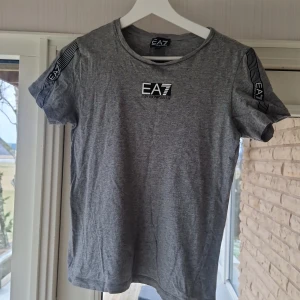 Grå t-shirt från Emporio Armani - Snygg grå t-shirt från Emporio Armani med EA7-logga på bröstet och ryggen. T-shirten har korta ärmar och en rund halsringning. Perfekt för en stilren och avslappnad look.