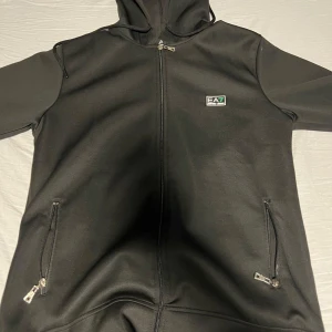 Svart hoodie från Emporio Armani - Säljer en stilren svart hoodie från Emporio Armani EA7. Den har dragkedja framtill och två praktiska fickor med dragkedjor. Perfekt för en casual look med en touch av lyx.
