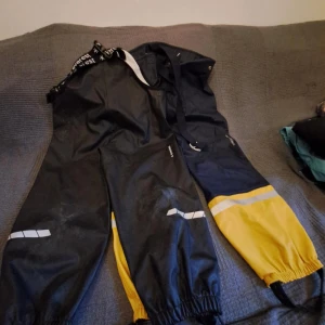  gula täckbyxor - Säljer ett par gula täckbyxor med elastiska muddar och reflexdetaljer. Perfekta för utomhusaktiviteter i kallt väder. Byxorna har justerbara hängslen för bättre passform.