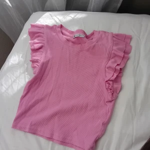 Rosa ribbad topp från Zara - Säljer en söt rosa ribbad topp från Zara med volangdetaljer vid ärmarna. Perfekt för en somrig look. Toppen är ärmlös och har en mjuk och bekväm passform. Lite liten i storleken 💗