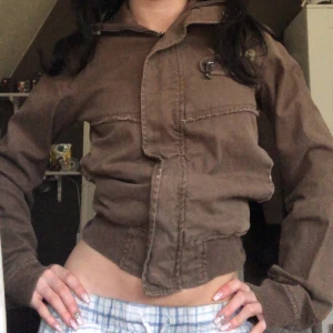 Brun y2k jacka med dragkedja - Säljer en brun jacka med dragkedja och ribbade muddar. Jackan har en croppad passform och långa ärmar. Perfekt för en avslappnad stil.