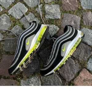 Nike Air Max 97 i svart och grå - Säljer ett par Nike Air Max 97 sneakers i svart och grå med neongula detaljer. Skorna har den ikoniska vågdesignen och synlig Air-enhet i sulan för extra komfort. Perfekta för en sportig look.