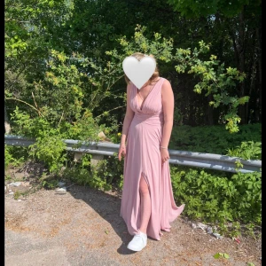 Ljusrosa lång balklänning - Elegant ljusrosa balklänning med v-ringad hals och draperad design, den har även insydd bh i innetyget. Klänningen har en slits framtill och är perfekt för sommarens festligheter. Den är ärmlös och har en mjuk och flödande passform.