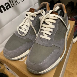 Grå sneakers från No Stress - Snygga grå sneakers från No Stress med vita snören och bruna detaljer. Skorna har en bekväm sula och är tillverkade i mocka och syntetmaterial. Perfekta för en avslappnad stil.