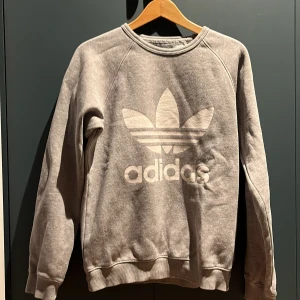 Grå sweatshirt från Adidas - Säljer en grå sweatshirt från Adidas med det klassiska trefoil-logotypen tryckt på framsidan. Tröjan har långa ärmar och en rund halsringning. Perfekt för en avslappnad stil.Lite sliten och nopprig på sina ställen men ändå bra skick👍Också vuxen storlek på denna