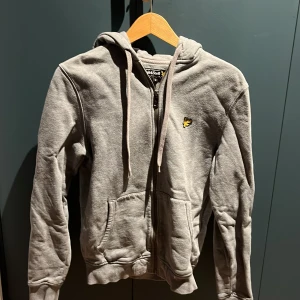 Grå hoodie från Lyle & Scott - Säljer en stilren grå hoodie från Lyle & Scott med dragkedja och huva. Tröjan har ett litet gult emblem på bröstet och är tillverkad i mjukt material. Perfekt för en avslappnad stil.Ganska urtvättad och blekt men ändå fint skick