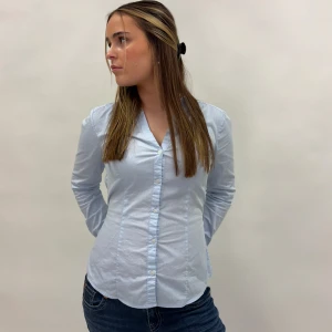 Ljusblå blus med knytband - Snygg ljusblå blus med knappar framtill och ett knytband i ryggen för en justerbar passform. Perfekt för en stilren look.