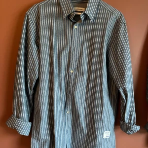 Randig skjorta från Jack & Jones - Snygg randig skjorta från Jack & Jones i blått och vitt. Skjortan har långa ärmar och knappar framtill. Perfekt för en stilren look med en touch av denimkänsla. Märkesdetalj med 'Brotherhood of Denim 1990' på framsidan.