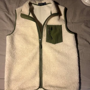 Vit fleeceväst från Polo Ralph Lauren - Säljer en snygg vit fleeceväst från Polo Ralph Lauren med gröna detaljer. Västen har en praktisk dragkedja framtill och en grön bröstficka med dragkedja och broderad logga. Perfekt för att hålla värmen med stil!