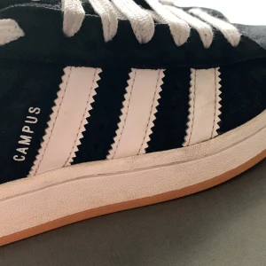 Svarta Adidas Campus sneakers - Snygga svarta Adidas Campus sneakers med vita detaljer och klassiska tre ränder på sidan. Skorna har en gummisula i brun färg och är tillverkade i mocka. Perfekta för en stilren look.