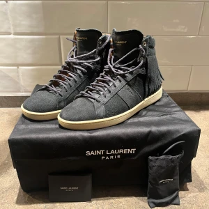  Saint Laurent skor - Snygga svarta sneakers från Saint Laurent i mocka med fransdetaljer på sidorna. Skorna har en klassisk snörning och en ljus sula som kontrasterar mot det mörka materialet. Perfekta för en stilren look.