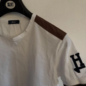 Vit och brun t-shirt från Herno - Snygg vit t-shirt från Herno med bruna axelpartier och ett stort H på ärmen. Perfekt för en stilren look. T-shirten har en klassisk rund hals och korta ärmar.