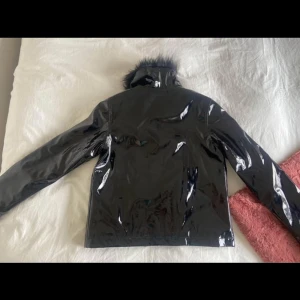 Svart glansig jacka med fuskpäls - Snygg svart glansig jacka med fuskpäls på kragen. Jackan har en stilren design med dragkedja framtill och långa ärmar. Perfekt för att ge en edgy touch till din outfit.
