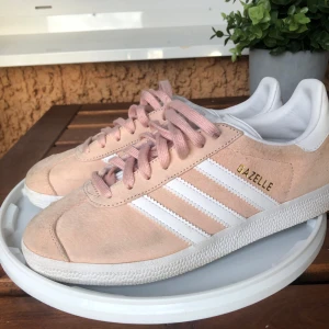 Adidas Gazelle i rosa mocka  - Snygga Adidas Gazelle sneakers i rosa mocka med vita detaljer och snörning. Perfekta för en stilren look med den klassiska tre-randiga designen på sidan och Gazelle-loggan i guld. Lite smutsigt med går att göra rent, passar storlek 37