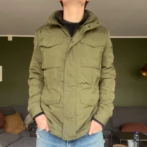 Olivgrön jacka från Bläck - Säljer en fieldjacka från Bläck med praktiska fickor och dragkedja. Jackan har en stilren design med långa ärmar och är perfekt för höst och vår. Skandinavisk stil med fokus på funktion och stil. Liten flaw på på armen, kan skicka bild om ni är intresserade! Hör av er vid frågor👍!