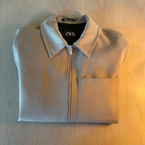 Beige jacka från Zara - Snygg beige jacka från Zara med dragkedja och en bröstficka. Jackan har en klassisk krage och är långärmad, perfekt för en stilren look. Passar bra till både vardag och mer uppklädda tillfällen.