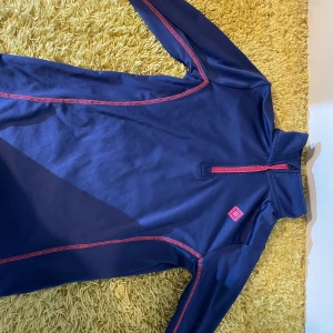 Mörkblå tröja från Crivit - Säljer en mörkblå tröja från Crivit med röda detaljer och dragkedja. Perfekt för milda vinterdagar. Tröjan har långa ärmar och är tillverkad i ett syntetmaterial som håller dig varm och bekväm.