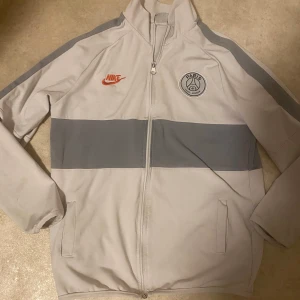 Psg jacka - Säljer en snygg vit och grå jacka från Nike i samarbete med Paris Saint-Germain. Jackan har en dragkedja framtill och långa ärmar. Den har en stor grå rand över bröstet och ryggen med texten 'PARIS' på baksidan. Perfekt för fotbollsfans! Storlek 160!!