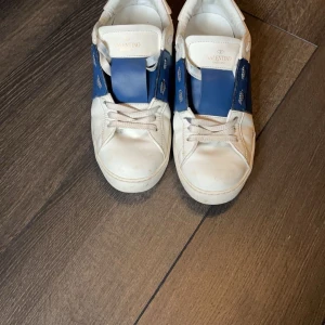 Valentino Garavani sneakers med blå detalj. Pris kan diskuteras - Snygga Valentino Garavani sneakers i vitt med en bred blå rem över snörningen. Skorna har en klassisk design med en modern twist och är tillverkade i Italien. Perfekta för att ge din outfit en lyxig touch.