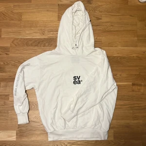 Vit  Svea Hoodie - Säljer en vit hoodie från Svea med tryck på bröstet och text längs ena ärmen. Den har en klassisk design med justerbar huva och ribbade muddar vid ärmslut och nederkant. Perfekt för en baggy stil.