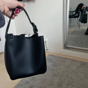 Svart handväska med axelrem - Snygg svart handväska i skinn med en justerbar axelrem. Väskan har en stilren design och en praktisk storlek för vardagsbruk. Perfekt för att bära dina nödvändigheter med stil.