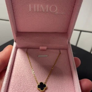 Guldhalsband med fyrklöver från HIMO - Elegant guldhalsband från HIMO med en fyrklöverformad berlock. Berlocken har en svart yta som ger en stilren kontrast till den gyllene kedjan. Perfekt för att lägga till en touch av lyx till din stil. Samt vattentåligt 