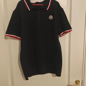 Svart pikétröja från Moncler - Säljer en stilren svart pikétröja från Moncler med röd och vit randig krage och ärmslut. Tröjan har en broderad logotyp på bröstet och knappar framtill. Perfekt för en casual look pris kan diskuteras 