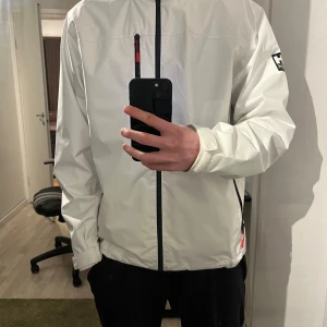 Vit vindjacka från Helly Hansen - Säljer en stilren vit vindjacka från Helly Hansen. Jackan har en dragkedja framtill och en praktisk bröstficka med dragkedja. Perfekt för blåsiga dagar och har en normal passform. Helly Hansen-logga på ärmen ger en sportig touch. Finns lite skavanker som att det är lite fläckar vid slutet av armarna men inget som man märker 