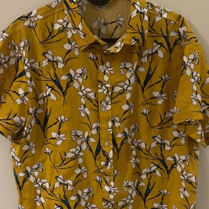 Gul blommig skjorta - Snygg gul skjorta med korta ärmar och ett blommigt mönster i vitt och blått. Perfekt för en somrig look med knappar framtill. Skjortan har en avslappnad passform och är tillverkad i ett lätt material.