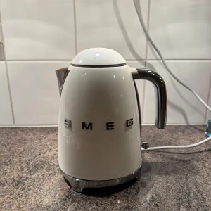 Vit vattenkokare från Smeg - Snygg och stilren vit vattenkokare från Smeg med en retro design. Den har en kromad pip och handtag samt tydliga bokstäver på sidan. Perfekt för att koka vatten snabbt och enkelt i köket. Välanvanvänd, men är fullt fungerande. Färgen har börjat lossna på två ställen, se bilder.