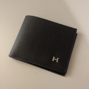 H-Plånbok - Snygg och stilren svart plånbok i skinn med en elegant H-detalj i metall på framsidan. Plånboken har flera fack för kort och en sektion för sedlar. Perfekt för den som vill ha en klassisk och tidlös accessoar.Det är bara att skriva vid fler frågor!😁