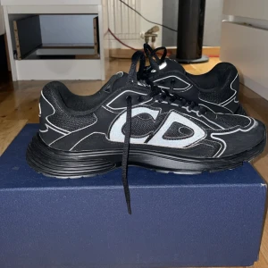 Svarta sneakers från Dior - Snygga svarta sneakers från Dior med vita detaljer och CD-logga på sidan. Skorna har snörning och en modern design som passar perfekt för en trendig look. Storlek 45/46 på dessa använda Max 5 gånger 