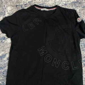 Svart t-shirt från Moncler - Snygg svart t-shirt från Moncler med ett diskret tryck på framsidan och ett broderat märke på ärmen. Tillverkad i Italien med högkvalitativt material. Perfekt för en stilren look.