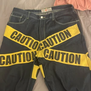Svarta jeans med varningstryck - Unika svarta jeans med iögonfallande gult 'CAUTION' tryck över benen. De har en loose passform och fransiga kanter vid bensluten för en edgy look. Perfekta för dig som vill sticka ut, jag är 173 så dom e lite långa med perfekta!😋😋😝