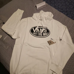 Vit hoodie från Vans - Säljer en snygg vit hoodie från Vans med det klassiska 'Off The Wall' trycket i svart på bröstet. Hoodien har en justerbar huva med snören och en stor magficka framtill. Perfekt för en avslappnad stil!
