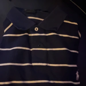 Randig pikétröja från Ralph Lauren - Säljer en klassisk mörkblå pikétröja från Ralph Lauren med vita ränder. Tröjan har en krage och knappar framtill, samt det ikoniska logotypbroderiet på bröstet. Perfekt för en stilren look.