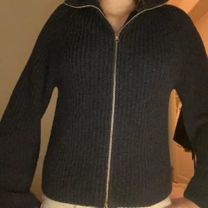 Svart stickad kofta med dragkedja - Säljer en svart stickad kofta med dragkedja och hög krage. Perfekt för kyliga dagar och ger en stilren look. Koftan har långa ärmar och en ribbad struktur som ger extra värme och komfort.