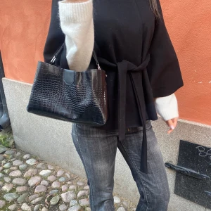 Svart handväska med krokodilmönster - Snygg svart handväska med krokodilmönster och korta handtag. Perfekt för att ge en stilren touch till din outfit. Väskan har en elegant design och är lätt att bära med sig.