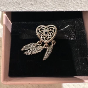 Silverfärgad berlock från Pandora - Säljer en vacker silverfärgad berlock från Pandora. Den har ett hjärtformat design med detaljerade utskärningar i form av en drömfångare. Perfekt för att ge ditt armband en unik touch.