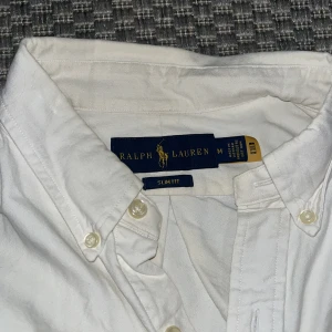 Vit skjorta från Ralph Lauren - Snygg vit skjorta från Ralph Lauren i slim fit. Skjortan har långa ärmar och klassiska knappar framtill. Den är broderad med den ikoniska loggan på bröstet. Perfekt för en stilren look. Dock är det en knapp som fattas i mitte av skjortan. Men det är bara att sy dit med de extra knapparna som vinns innanför skjortan! Fråga gärna ifall du funderar över något!😽💗