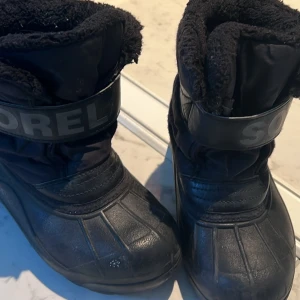 Svarta vinterstövlar från Sorel - Säljer ett par svarta vinterstövlar från Sorel. De har en robust design med vadderad insida för extra värme. Stövlarna har en bred rem med Sorel-logga och en slitstark sula för bra grepp på hala underlag.  Storlek 30