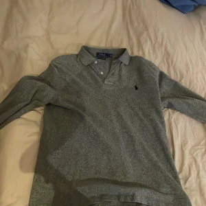 Grå pikétröja från Ralph Lauren - Säljer en stilren grå pikétröja från Ralph Lauren i custom slim fit. Tröjan har en klassisk krage och knappar framtill. Perfekt för en avslappnad men ändå snygg look.