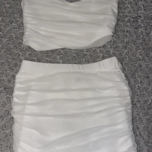 Vit croppad topp och kjol från Shein - Säljer en stilren vit croppad topp och kjol från Shein. Toppen har en omlott-design och är ärmlös, perfekt för varma dagar eller sommarfester. Den är tillverkad i ett mjukt och bekvämt material.